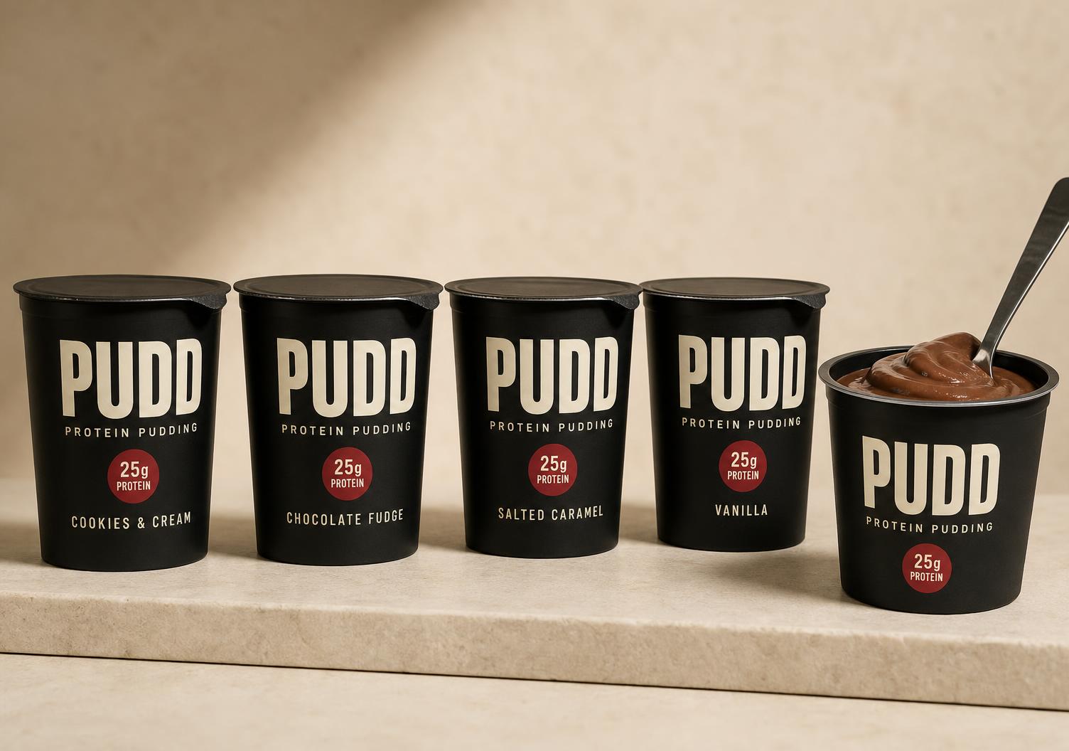 Four matte black PUDD Protein Pudding tubs in Cookies & Cream, Chocolate Fudge, Salted Caramel and Vanilla with one open tub of chocolate pudding